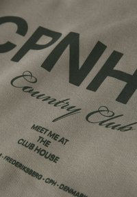 Bavlněná látka v světle šedé barvě obsahuje silný černý text s nápisem "CPNH Country Club" a menším textem níže, který zdůrazňuje pozvání do klubovny.