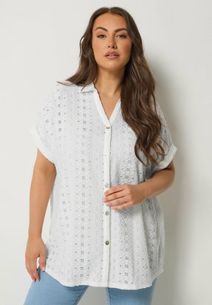 Kobieta o długich brązowych włosach ubrana w białą, krótkorękawową bluzkę z guzikami wykonanymi z ażurowej tkaniny oraz jasnoniebieskie dżinsy, stojąca na tle jednolitego tła.