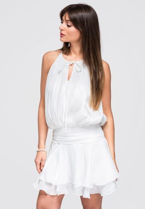 Femme aux longs cheveux bruns portant une robe blanche sans manches avec un décolleté en trou de serrure et une jupe à volants, regardant vers sa gauche.