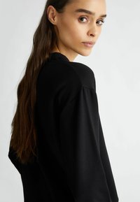 Felpa nera con colletto rotondo, texture morbida e maniche lunghe. Modella con lunghi capelli, vista di lato, che mette in evidenza il design.