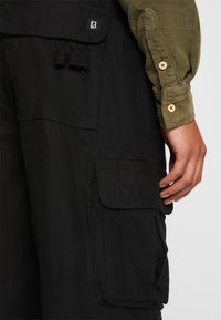 Černé cargo kalhoty mají několik bočních kapes, texturovanou látku a nastavitelné pásko. Doprovodná košile je olivově zelená s knoflíkovými akcenty.