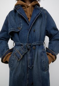 Manteau en jean avec une taille ceinturée, un grand col et une fermeture à boutons. Il présente une couleur bleue délavée et des poches latérales, superposé à un sweat à capuche marron.