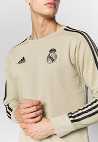 Giovane uomo che indossa una felpa Adidas beige con strisce nere sulle maniche e il logo del Real Madrid, mani incrociate in vita.