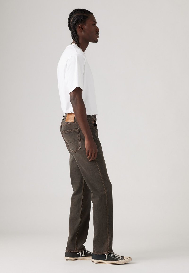 Weißes Kurzarm-T-Shirt kombiniert mit dunkelbraunen Jeans. Die Jeans haben ein Lederpatch an der Taille und werden durch schwarze Sneakers ergänzt.