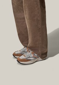 Baskets athlétiques grises et brunes avec des accents en cuir et en daim, dotées d'une semelle texturée et de détails orange, associées à un pantalon marron.