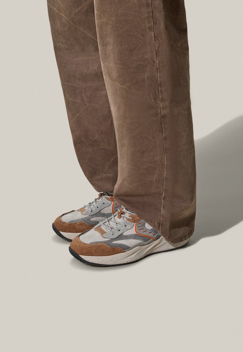 Baskets athlétiques grises et brunes avec des accents en cuir et en daim, dotées d'une semelle texturée et de détails orange, associées à un pantalon marron.