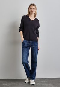 Marineblauer V-Ausschnitt-Pullover mit geripptem Muster, kombiniert mit blauen Jeans im geraden Schnitt. Das Model steht mit einer Hand in der Tasche und trägt hellfarbene Sneakers.