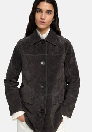 Kobieta ubrana w ciemnoszarą zamszową kurtkę zapinaną na guziki, na białym swetrze z dzianiny, z rękami w kieszeniach, zwrócona przodem na jednolitym tle.