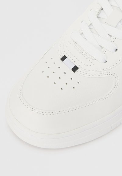 Sapatilha branca de couro com textura suave, bico redondo, detalhes perfurados, cadarços brancos e um pequeno acento preto perto dos ilhós.