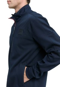 Homme portant une veste matelassée bleu marine avec un col haut, des poches latérales et une fermeture éclair, la main reposant dans la poche, faisant face à gauche.