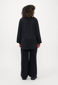 Zwarte top met een gespikkeld patroon, lange mouwen en een losse pasvorm, gecombineerd met brede zwarte broeken en chunky zwarte schoenen.