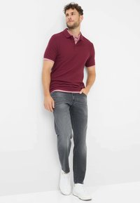 Polo bordeaux a maniche corte con un colletto a due bottoni, abbinato a jeans grigi e sneaker bianche, appoggiato a un muro.