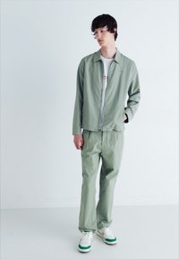 Jachetă verde deschis și pantaloni asortați dintr-un material ușor. Tricou alb pe dedesubt. Încălțăminte: adidași albi cu accente verzi.