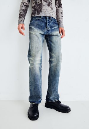 Ausgebleichte blaue Jeans im geraden Schnitt mit leichter Abriebstelle, kombiniert mit schwarzen klobigen Schuhen. Der Stoff zeigt Anzeichen von Abnutzung und leichtem Faltenwurf.