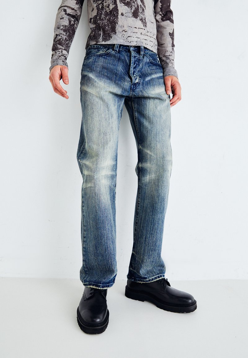 Faded blue jeans with a straight fit and light distressing, paired with black chunky shoes. Fabric shows signs of wear and whiskering.