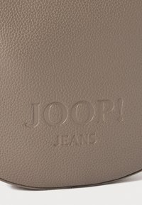 Šedá texturovaná kůže s vyraženým logem "JOOP! JEANS" uprostřed. Kulatý tvar s hladkým povrchem a jemným zrnitým vzorem.