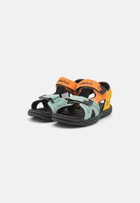 Urheilusandaalit, joissa on vihreät ja oranssit synteettiset hihnat, mustat kumipohjat ja säädettävät Velcro-kiinnitykset turvalliseen istuvuuteen.