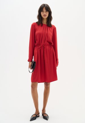 Rotes langärmeliges Kleid mit Kordelzug in der Taille, plissiertem Vorderteil und weichem Stoff. Getragen mit schwarzenFlats und einer kleinen schwarzen Handtasche.