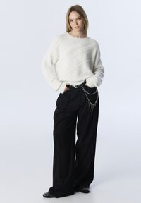 Pull en maille texturée blanche avec un motif en côtes diagonales. Pantalon noir à jambes larges avec une ceinture en chaîne et des accents cloutés sur les chaussures.