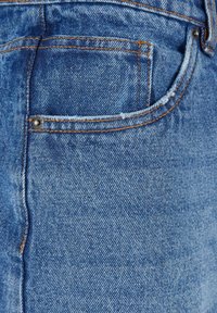 Blå denimtyg med en strukturerad yta, som har en bakficka med sömnadsdetaljer och en metallnitar, allt i en klassisk rak design.