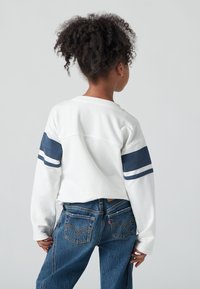 Lapsi, jolla on kiharat hiukset, valkoisessa pitkähihapuserossa, jossa on sinisiä raitoja, ja siniset farkut, nähtynä takaa yksinkertaista taustaa vasten.