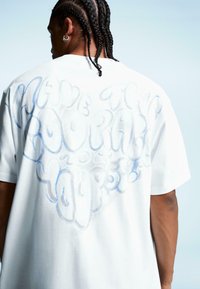 Tricou alb din bumbac cu grafica mare de text în stil bubble de culoare albastră și argintie pe spate, care citește "Fii îndrăzneț să fii tu însuți."