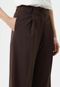 Pantalon marron sur mesure avec plis, fermeture par bouton et poches latérales. Tissu lisse avec un léger éclat et coupe droite.