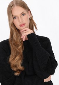 Sort strikket sweater med struktureret mønster og ribstrikkede manchetter. Modellen har langt bølget hår og holder en hånd nær sin hage.