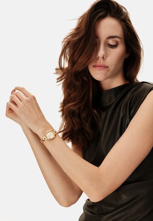 Femme aux longs cheveux bruns portant un haut noir sans manches ajustant une montre-bracelet à maillons dorés sur son poignet gauche.