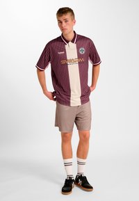 Koszulka w paski bordo i kremowa z krótkimi rękawami, zestawiona z jasnoszarymi szortami. Na koszulce znajduje się logo "SWARM" oraz dodatkowe akcenty. Czarne trampki i skarpetki w paski.