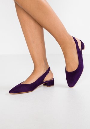 Kvinde iført lilla ruskind slingback-sko med lave blokhæle, stående på en lys overflade mod en hvid baggrund.