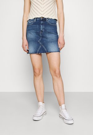 Woman wearing a blue denim mini skirt with frayed hem, a beige knit sleeveless top, and white high-top sneakers, standing on a gray floor.