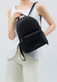 Musta nahkainen reppu, jossa on pyöristetty muoto, kultaketju, etutasku ja säädettävät hihnat. Teksturoitu pinta ja minimalistinen muotoilu.