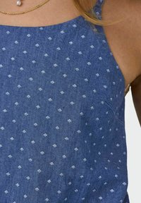 Blauwe stof met een patroon van kleine witte diamanten. Heeft dunne banden en een scoophalslijn, gemaakt van lichte, gestructureerde materialen.