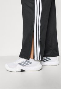 Črne športne hlače z belimi stranskimi črtami in z zadrgami na robu, kombinirane z belimi športnimi čevlji z sivimi poudarki.