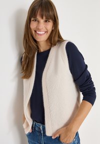 Beige ull utan ärmar väst med en mjuk textur, öppen framtill, bärs över en marinblå långärmad topp. Modellen poserar med händerna i fickorna.