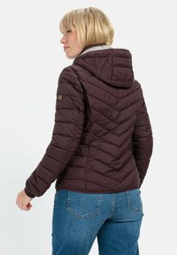 Braune Pufferjacke mit Kapuze, mit einem Chevron-Muster, Quilt-Textur und gerippten Bündchen. Kombiniert mit blauen Jeans.