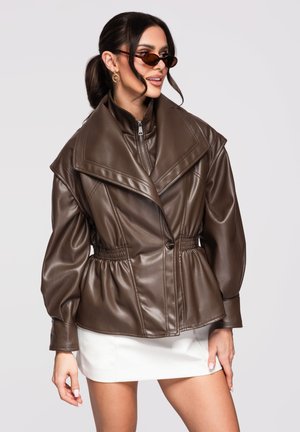 Donna che indossa una giacca di pelle marrone oversize con collo ampio, minigonna bianca, occhiali da sole e orecchini a cerchio dorati, in piedi davanti a uno sfondo semplice.