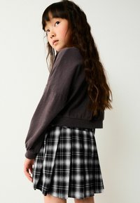 Brauner Crop-Sweatshirt mit langen Ärmeln, kombiniert mit einem schwarz-weiß karierten, gefältelten Rock. Strukturierter Stoff, sichtbare Falten, lässiges Design.