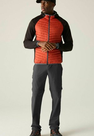 Man met een zwarte pet, een rood-zwarte doorgestikte jas, donkergrijze broek en zwarte wandelschoenen staat met gevouwen handen.