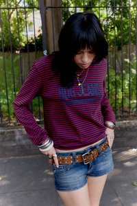 Cămașă cu mânecă lungă cu dungi roșii și bleumarin, combinată cu pantaloni scurți din denim și o curea maro cu ținte. Accesorii includ brățări argintii și coliere suprapuse.