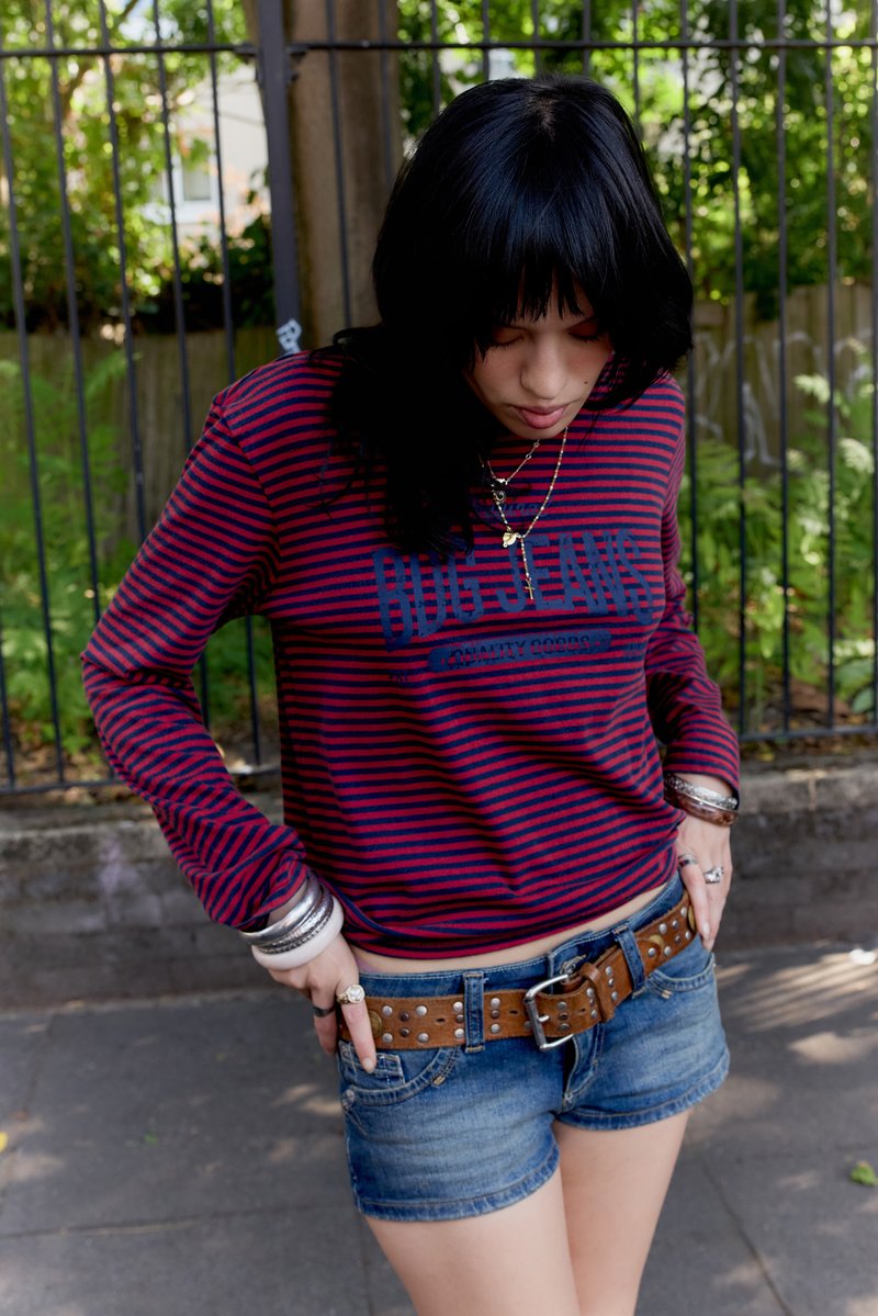 Cămașă cu mânecă lungă cu dungi roșii și bleumarin, combinată cu pantaloni scurți din denim și o curea maro cu ținte. Accesorii includ brățări argintii și coliere suprapuse.