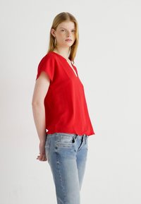 Blusa rossa a maniche corte con scollo a V e tessuto testurizzato, abbinata a jeans azzurri chiari. La blusa ha una vestibilità rilassata e spacchi laterali.
