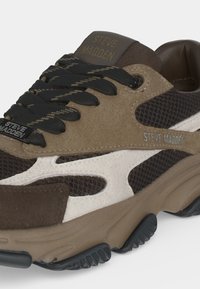 Кафяво и черно кецове Steve Madden с черни връзки с шарка, бели странични акценти и масивна подметка, видима от страничен ъгъл.
