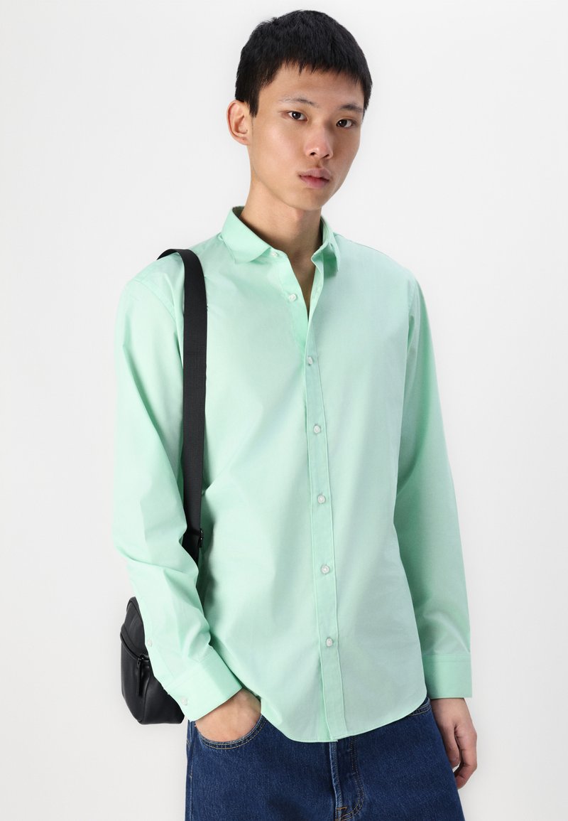 Camisa de botón verde menta con mangas largas, fabricada en suave algodón, con un bolsillo en el pecho, combinada con jeans de mezclilla azules y un bolso negro de hombro.