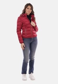 Giacca isolante rossa con cappuccio, chiusura con zip e due tasche laterali, abbinata a jeans blu e sneakers bianche. Texture liscia.