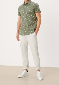 Camisa de manga corta verde oliva con un patrón de palmeras blancas, combinada con pantalones claros beige arremangados y zapatillas blancas.
