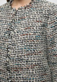 Jachetă tweed multicoloră cu margine despicată, având fire negre, albe, roșii și albastre. Țesătură texturată, împletitură strânsă, cu o formă structurată.