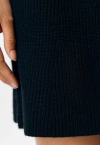 Nahaufnahme von geripptem schwarzem Strickstoff mit einer strukturierten Oberfläche und einem Saum. Eine Hand berührt das Material und betont dessen Weichheit.