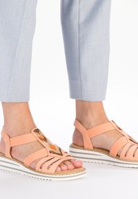 Sandales de couleur pêche avec un design à lanières, présentant un accent en perles et une semelle rayée blanche. Matériau doux avec une finition texturée.